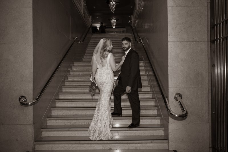 Bride in a detailed gown and veil touches groom's tie on grand staircase, illuminated by chandeliers.