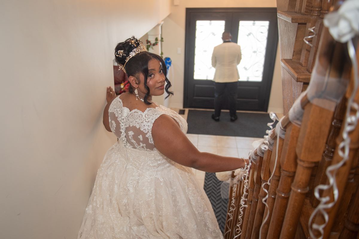 A bride in an ornate white gown with a tiara pauses on a staircase, looking back over her shoulder with a gentle smile.