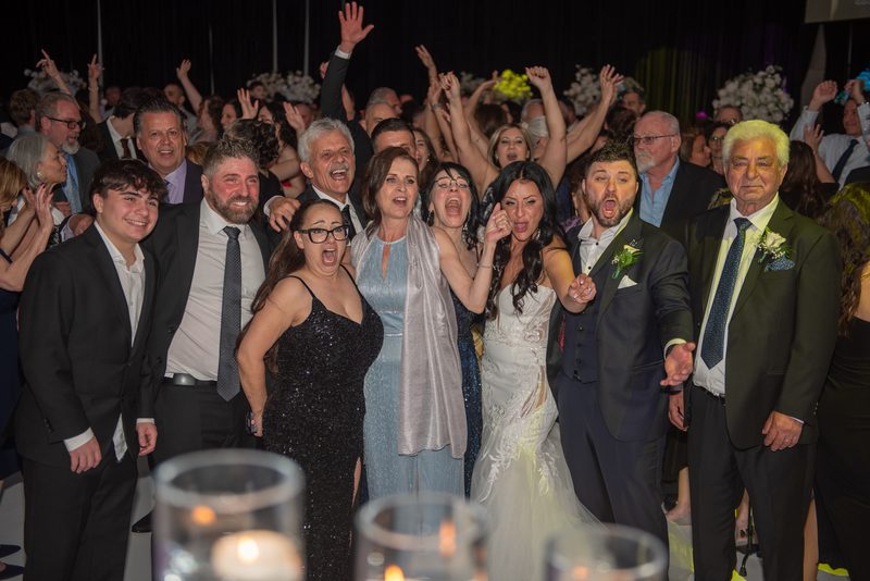 Sarah and Alessandro surrounded by cheering guests at their wedding reception, everyone raising hands in joy.