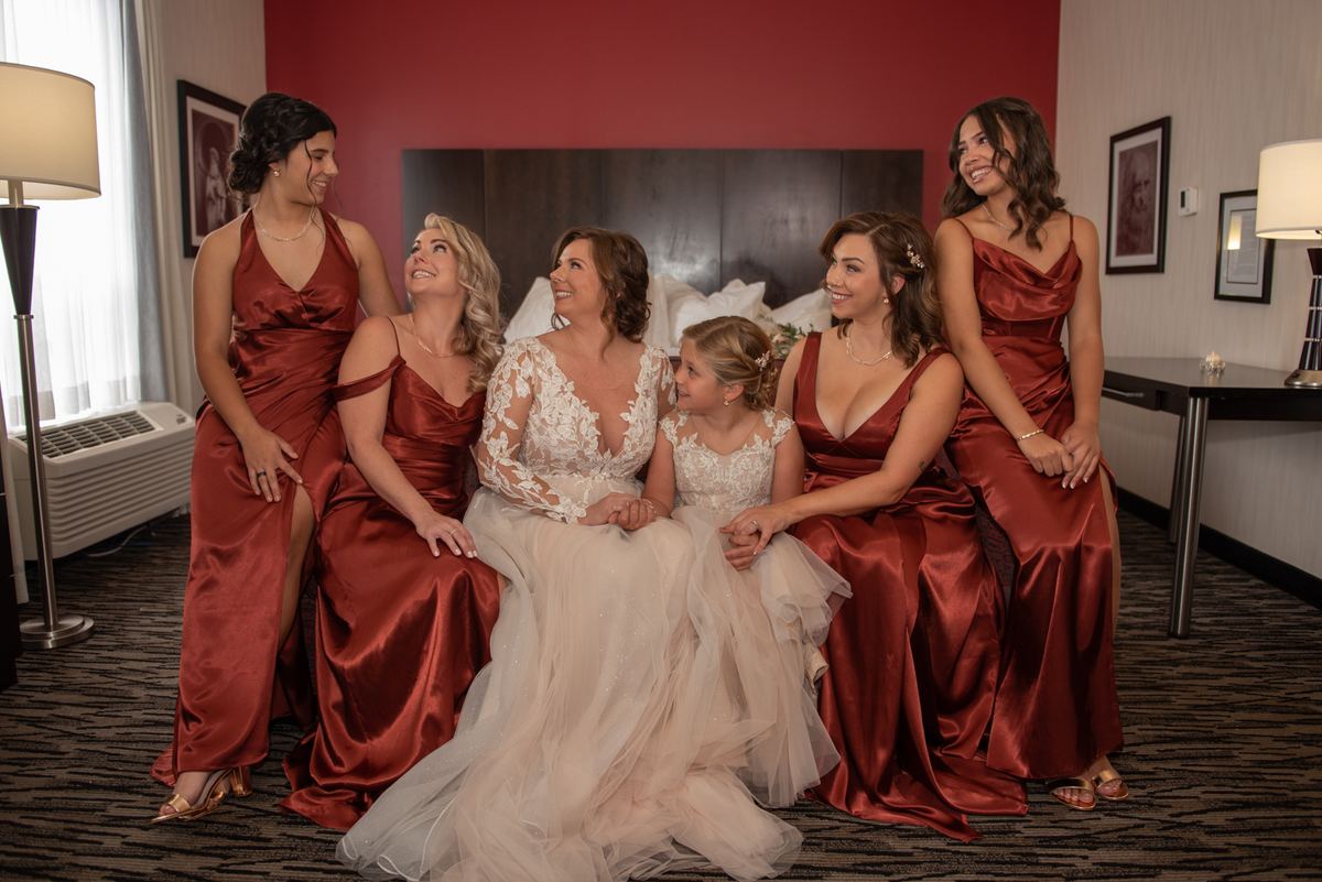 A bride and her bridesmaids in a hotel room, smiling and looking upward, illuminated by soft light from lamps and a window.