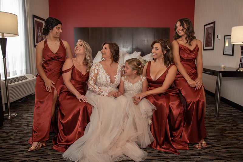 A bride and her bridesmaids in a hotel room, smiling and looking upward, illuminated by soft light from lamps and a window.