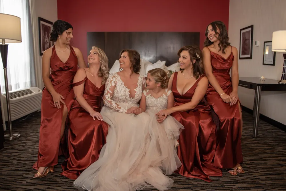 A bride and her bridesmaids in a hotel room, smiling and looking upward, illuminated by soft light from lamps and a window.