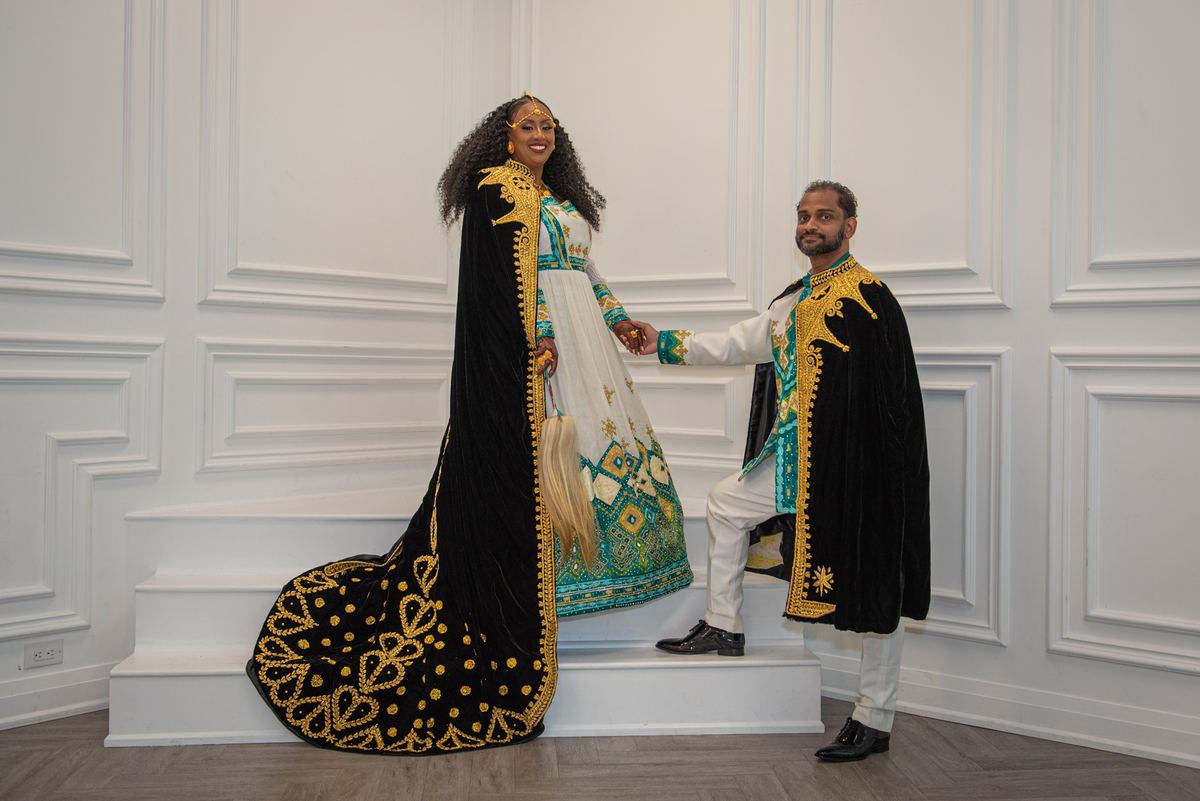 A couple in ornate black velvet and gold-embroidered attire stand on white stairs, smiling against a paneled wall.