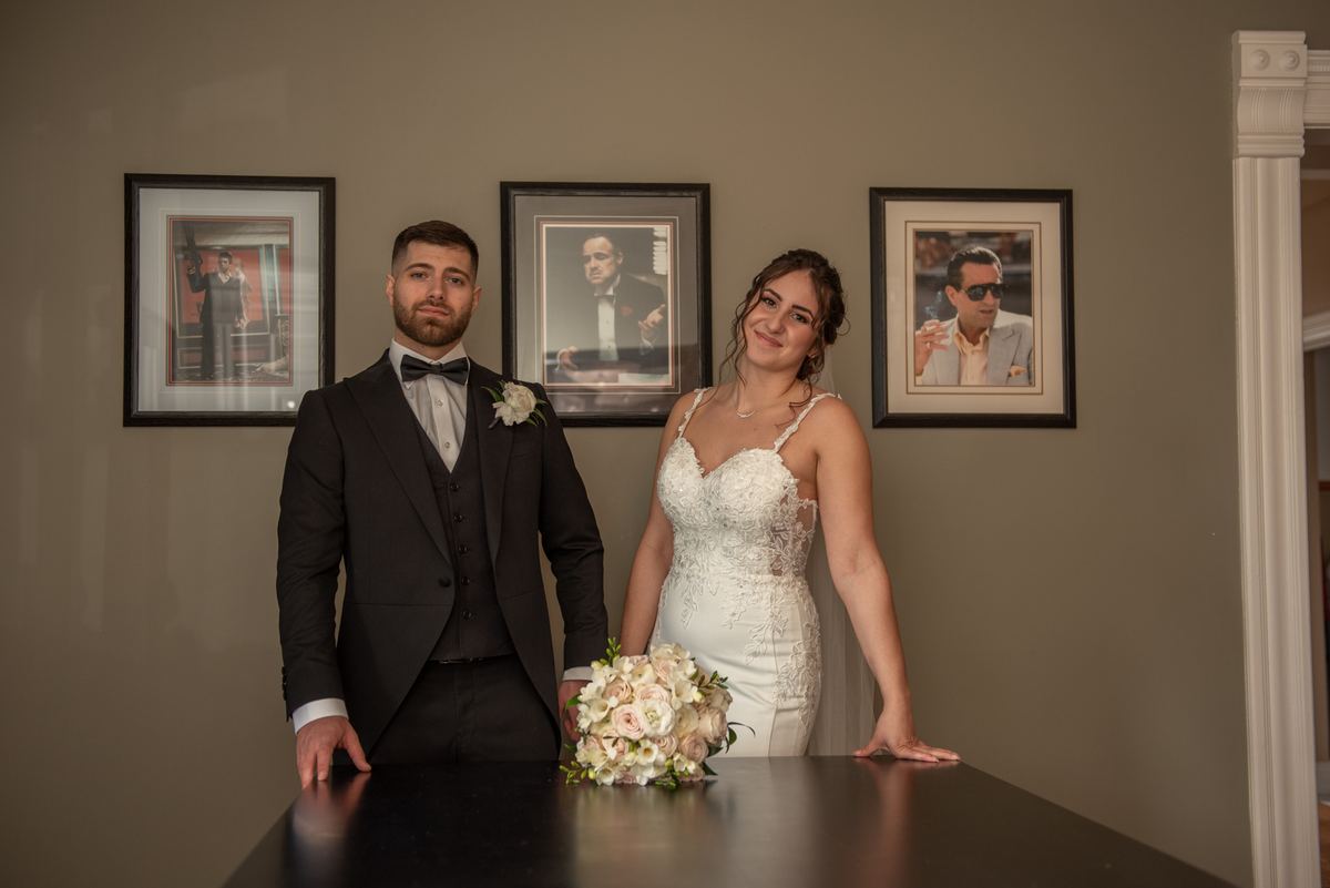 A bride and groom pose in front of framed mobster images, contrasting their wedding attire with the crime theme decor.
