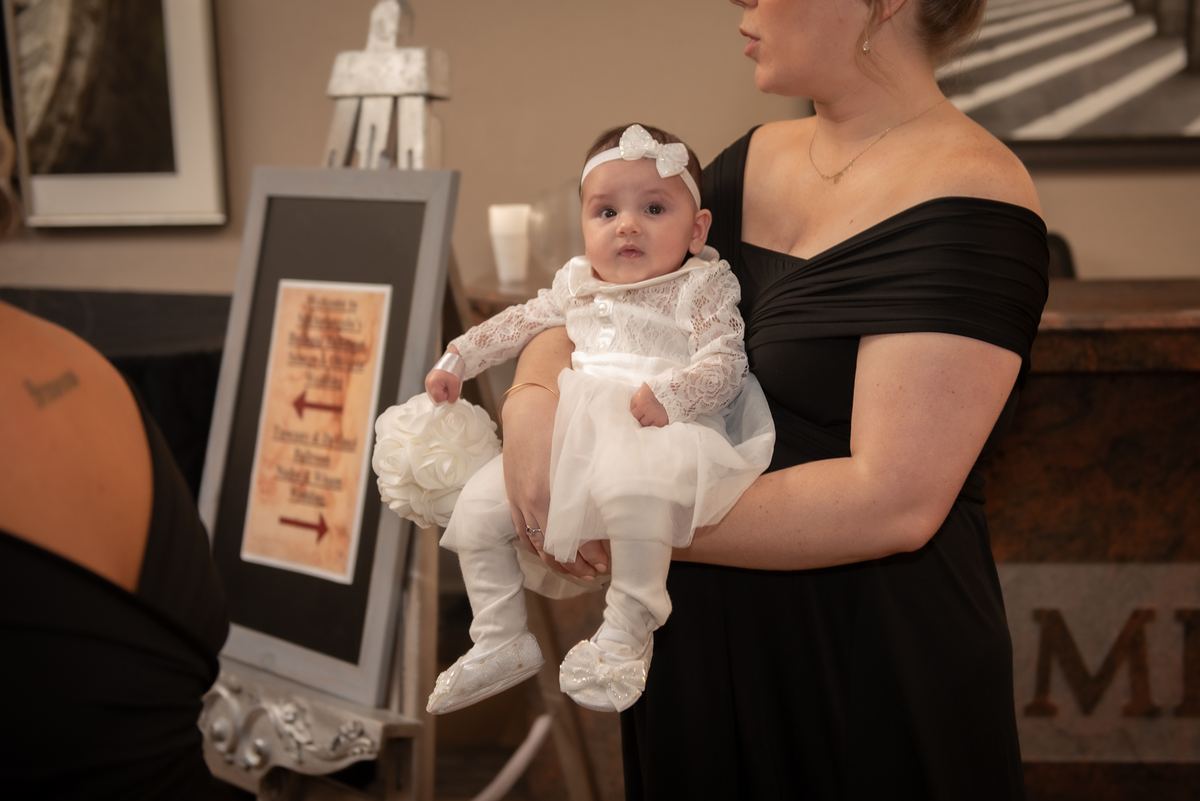 A woman in a black dress holds a baby dressed formally, with signage suggesting a celebration in the background.
