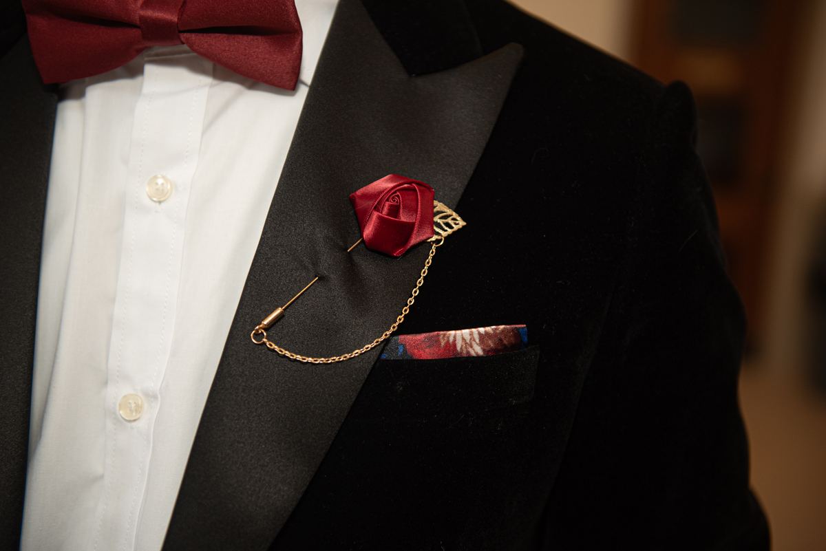 A formally dressed man in a tuxedo with a burgundy bow tie and satin rose boutonnière, at an indoor evening event.