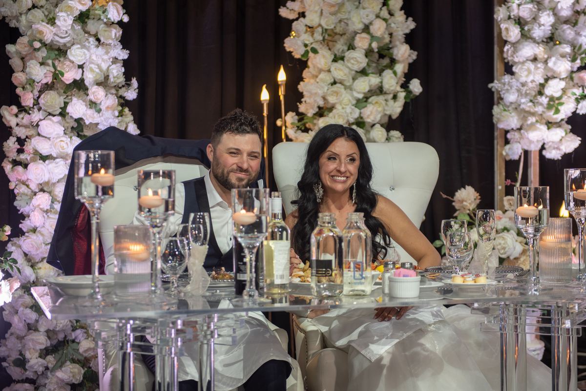 A smiling couple sits at a lavishly decorated table with tall floral arrangements and candlelight during a wedding reception.