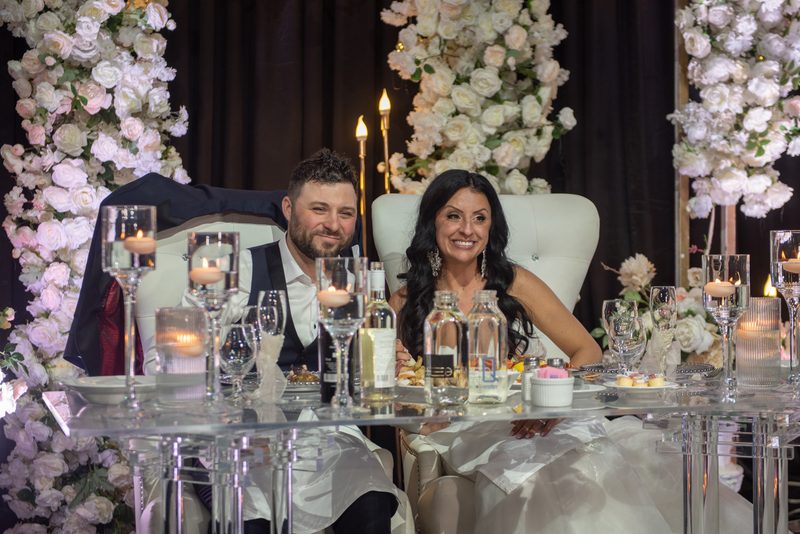A smiling couple sits at a lavishly decorated table with tall floral arrangements and candlelight during a wedding reception.