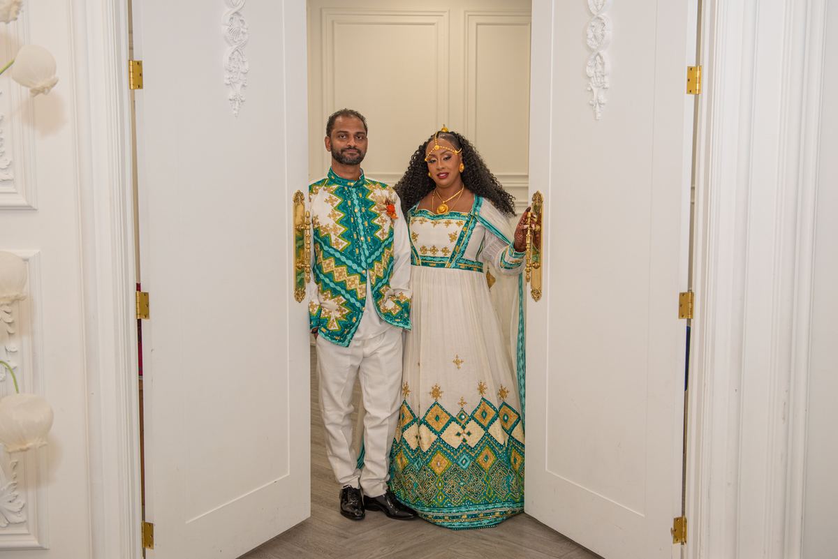 A couple in vibrant traditional attire stands ceremonially in an ornate doorway, illuminated by warm light.
