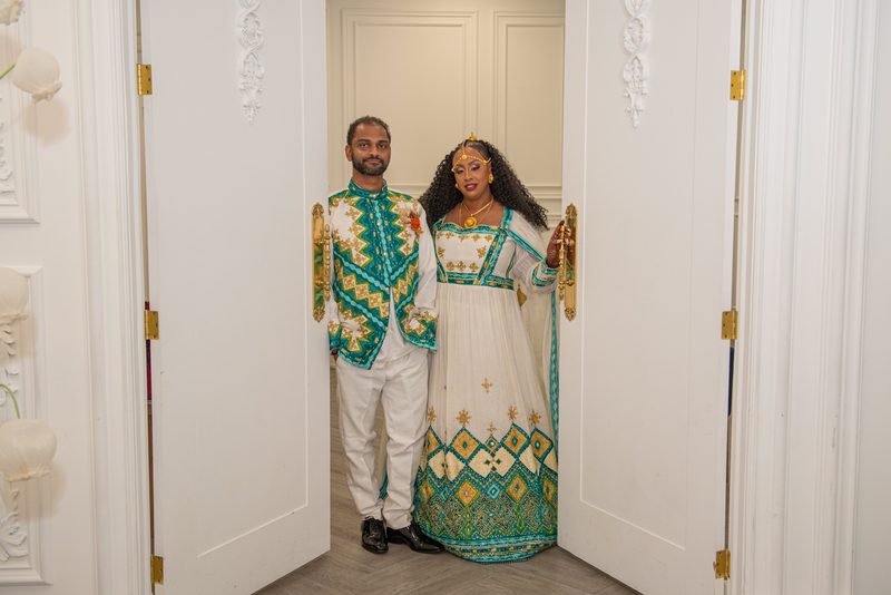 A couple in vibrant traditional attire stands ceremonially in an ornate doorway, illuminated by warm light.