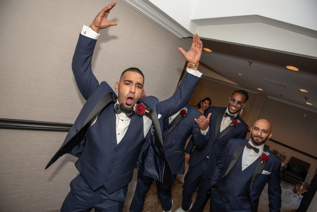 A groom and his groomsmen celebrate in matching navy tuxedos with raised arms and smiles, under warm lights.