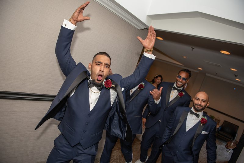 A groom and his groomsmen celebrate in matching navy tuxedos with raised arms and smiles, under warm lights.