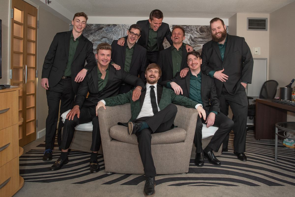 Seven men in black suits with green shirts pose joyfully around an armchair in a hotel room, celebrating.