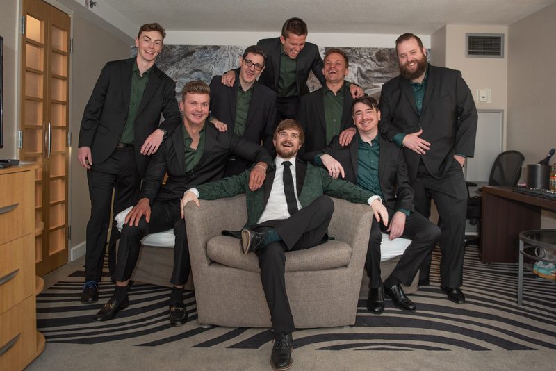 Seven men in black suits with green shirts pose joyfully around an armchair in a hotel room, celebrating.