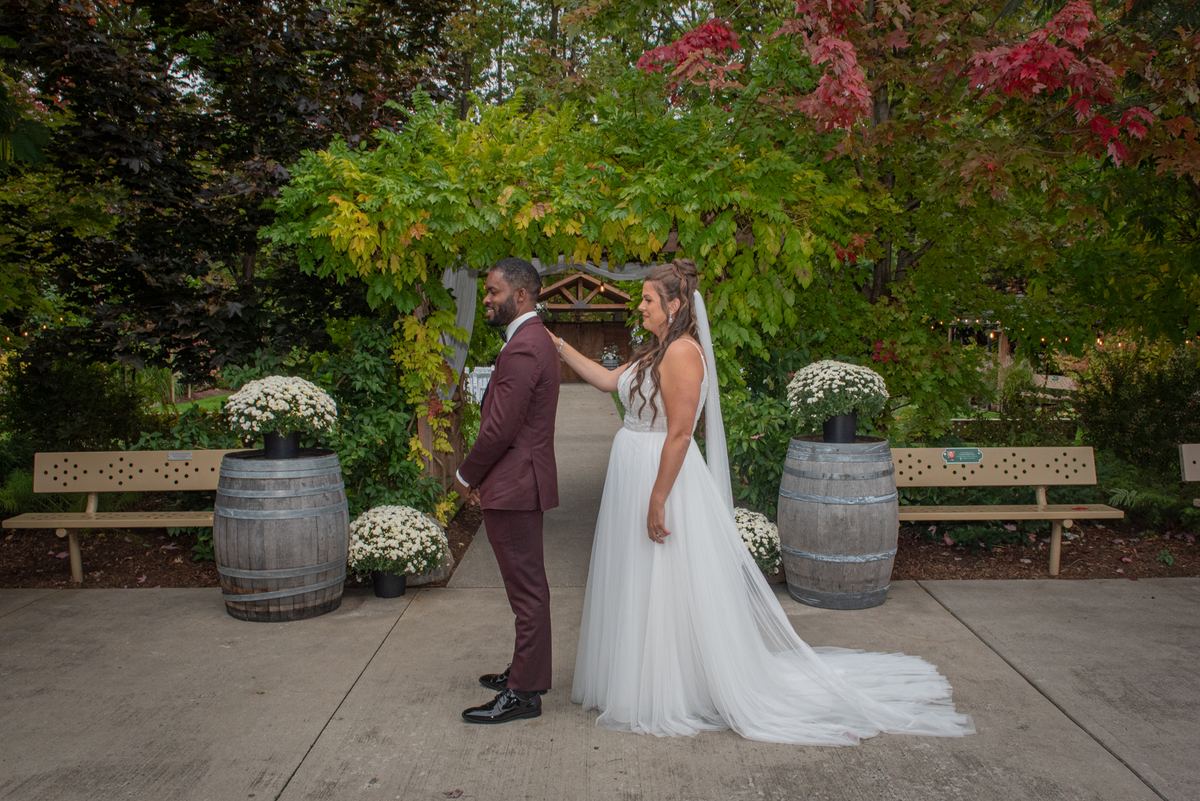 Bride in white gown and veil stands with groom in garden surrounded by autumn foliage and barrel planters.