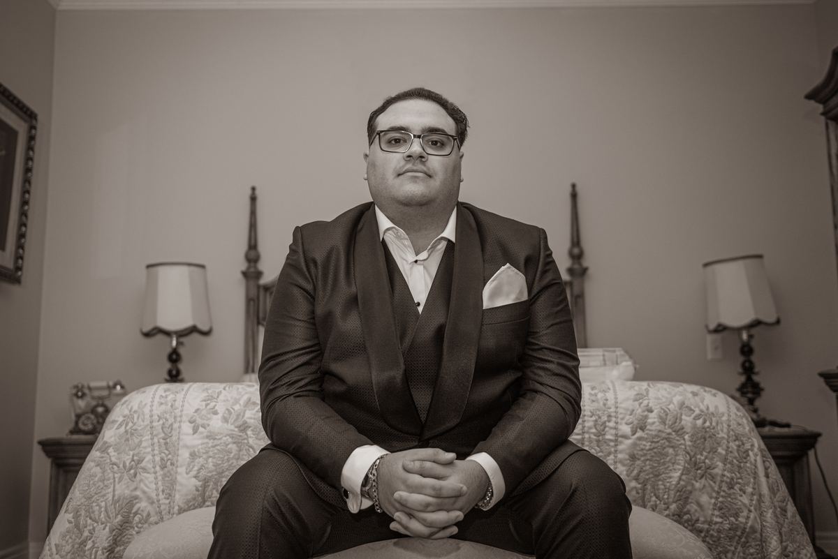 A groom sits formally on an ornate bed, hands clasped, gazing directly at the camera in a vintage bedroom.
