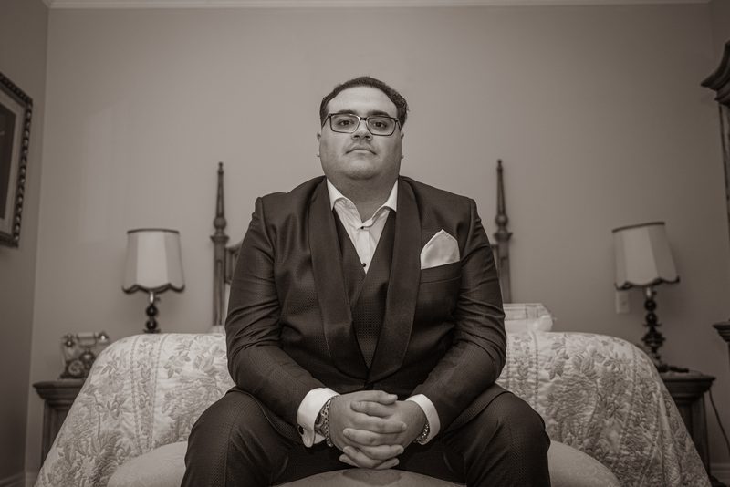 A groom sits formally on an ornate bed, hands clasped, gazing directly at the camera in a vintage bedroom.