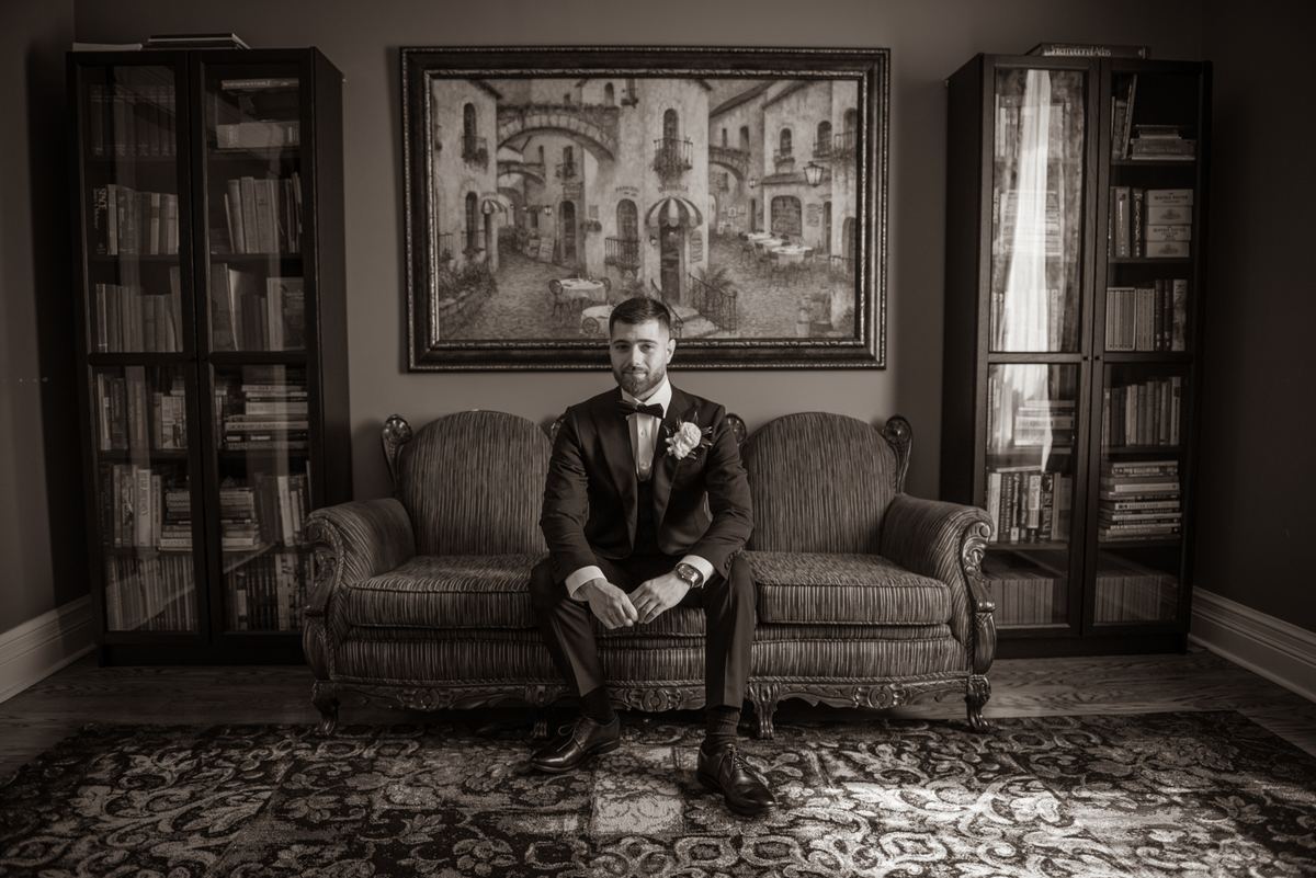 A groom in a tuxedo sits composedly on an ornate sofa, illuminated by a shaft of light from a window.