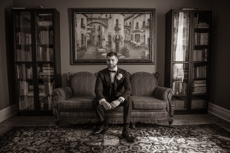 A groom in a tuxedo sits composedly on an ornate sofa, illuminated by a shaft of light from a window.