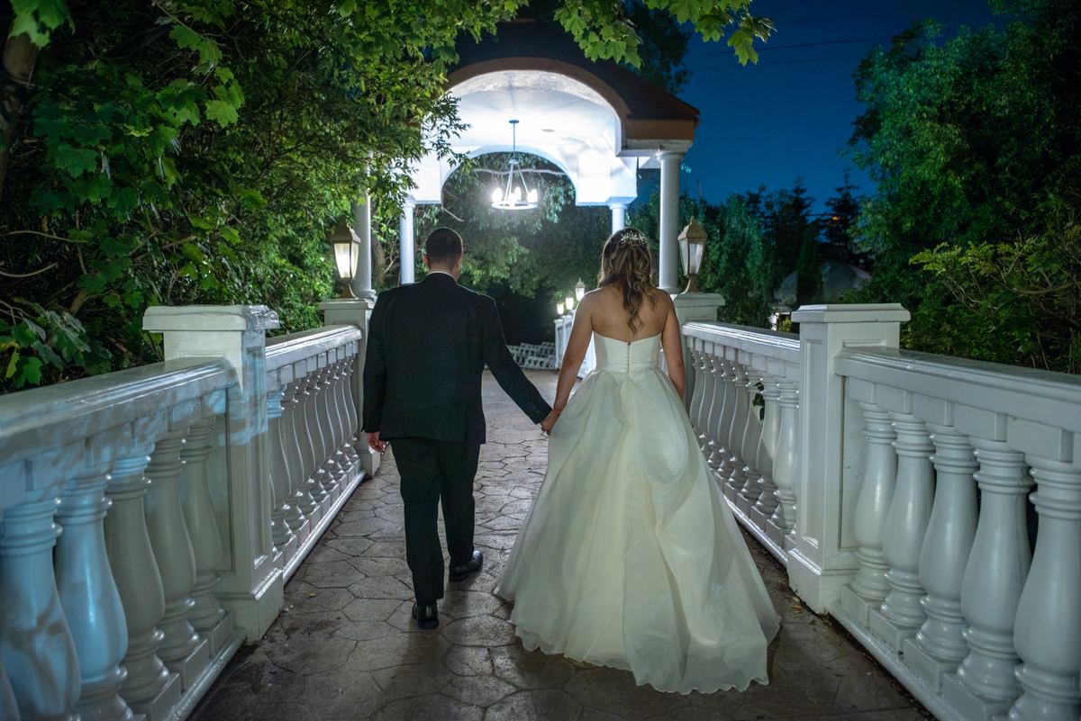 A bride and groom walk across a stone bridge at night, heading toward a lit gazebo surrounded by dark foliage.