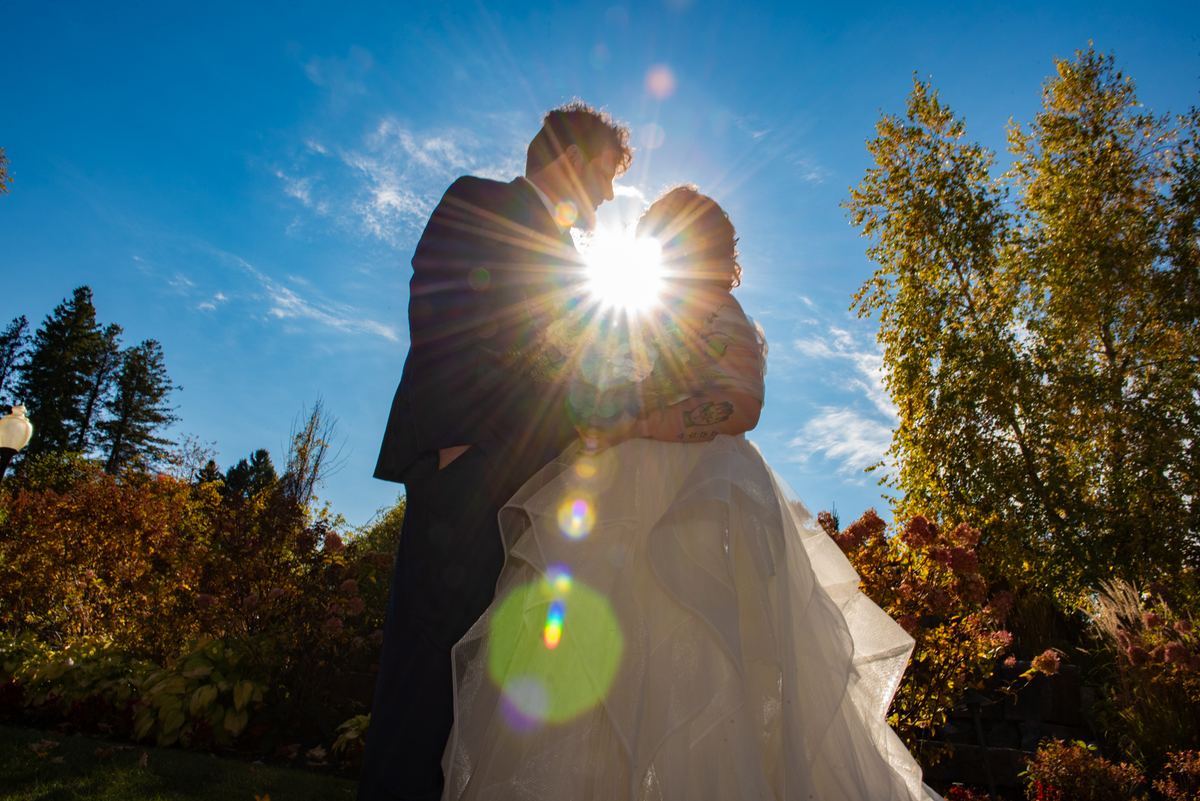 A bride and groom embrace in a garden, silhouetted by sunlight with lens flares, surrounded by autumn foliage.