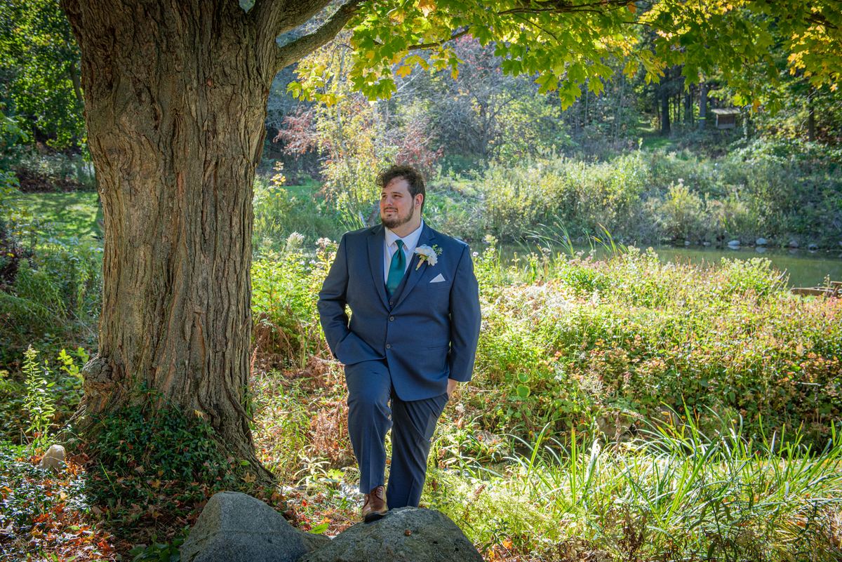 A groom in a navy suit stands beside a large tree, smiling as sunlight filters through autumn leaves.