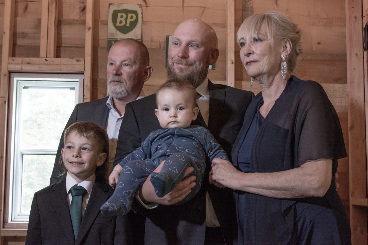 A family in formal attire gathers inside a rustic wooden building with soft natural light, holding a baby.