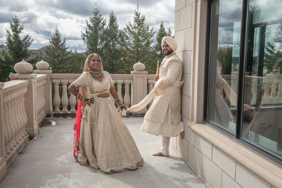 A smiling bride in traditional South Asian attire and her groom pose happily on a rooftop terrace under an overcast sky.