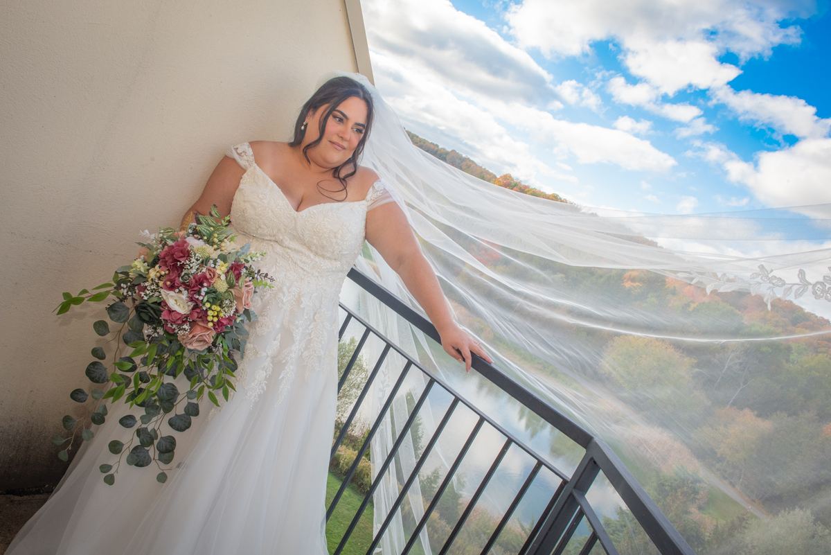 A bride in a lace gown stands on a balcony, her veil fluttering in the breeze over autumn hills and blue sky.