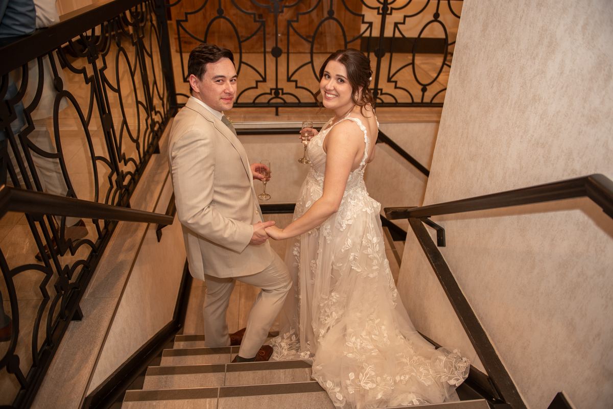 A bride in lace and a groom in a suit descend a grand staircase with an ornate iron railing, holding champagne flutes.
