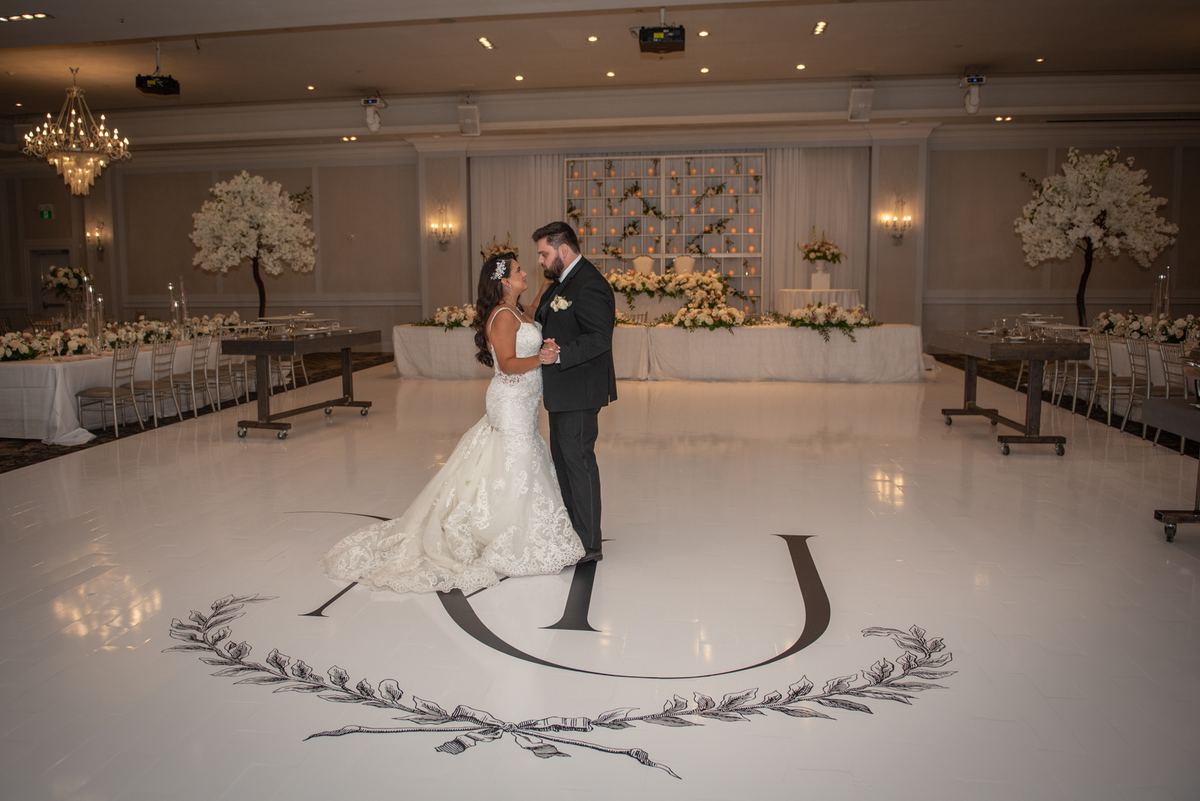 A bride and groom dance intimately on a monogrammed floor in an elegantly lit ballroom, surrounded by floral arrangements.