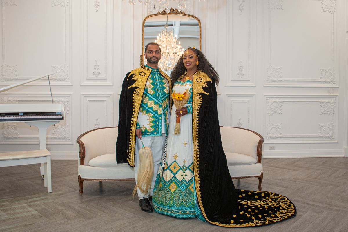 A couple in ornate traditional attire stands smiling in an opulent room with a chandelier and grand piano.