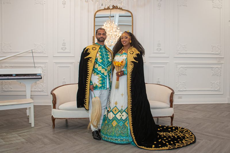 A couple in ornate traditional attire stands smiling in an opulent room with a chandelier and grand piano.