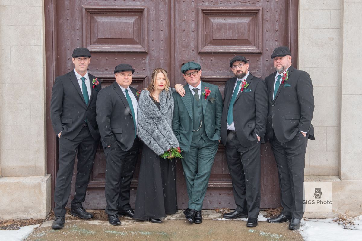 A bride in a black dress and fur stole stands with five groomsmen in dark suits by an ornate wooden door on a snowy day.