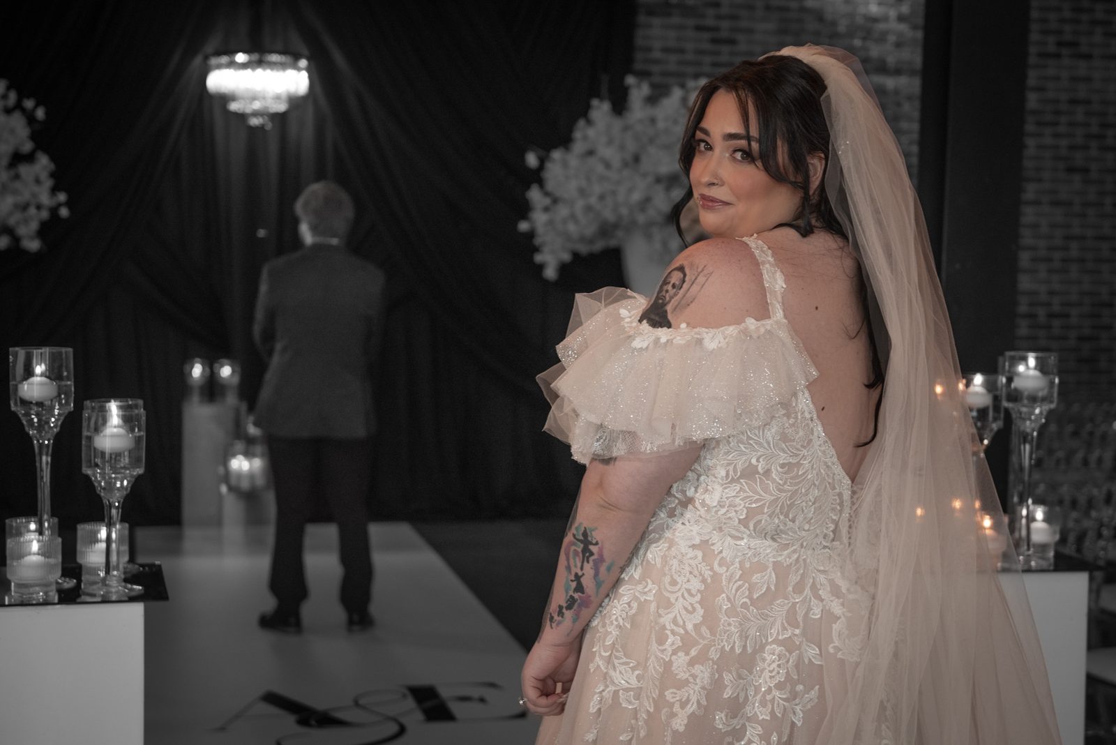 A bride in a lace gown turns to look at the camera while her groom stands behind her in a dimly lit aisle with candles.