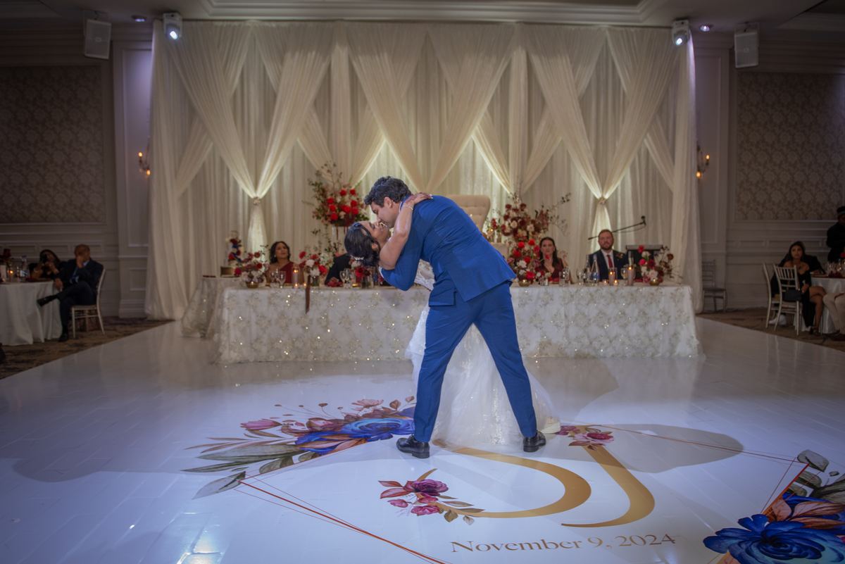 A bride and groom dance on a decorated floor, surrounded by white curtains and seated guests at an elegant table.