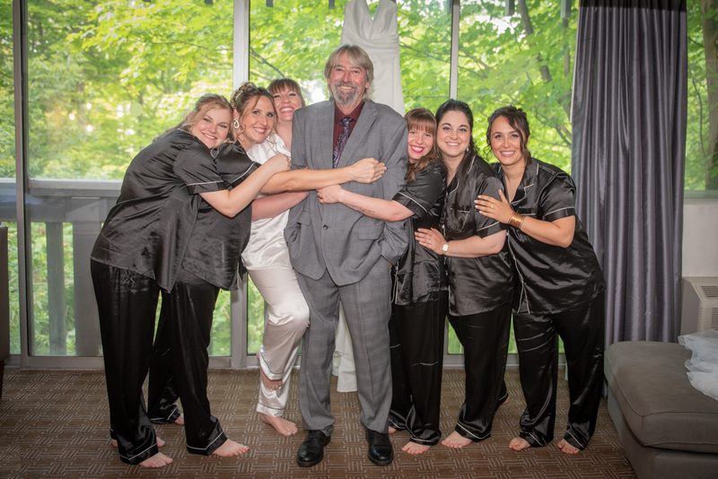 Samantha is surrounded by her bridesmaids and a smiling older man in an indoor setting, all dressed in satin pajamas.