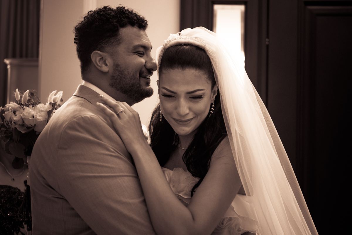 A bride with closed eyes smiles as her groom holds her close in a sepia-toned embrace lit by soft light from one side.