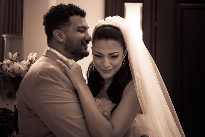 A bride with closed eyes smiles as her groom holds her close in a sepia-toned embrace lit by soft light from one side.