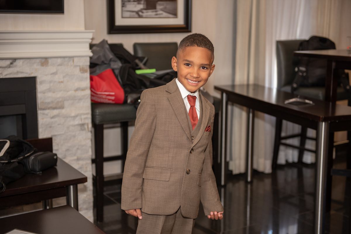 A young boy in a brown suit, white shirt, and red tie smiles in a hotel lobby with luggage and tables nearby.