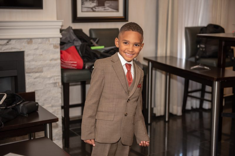 A young boy in a brown suit, white shirt, and red tie smiles in a hotel lobby with luggage and tables nearby.