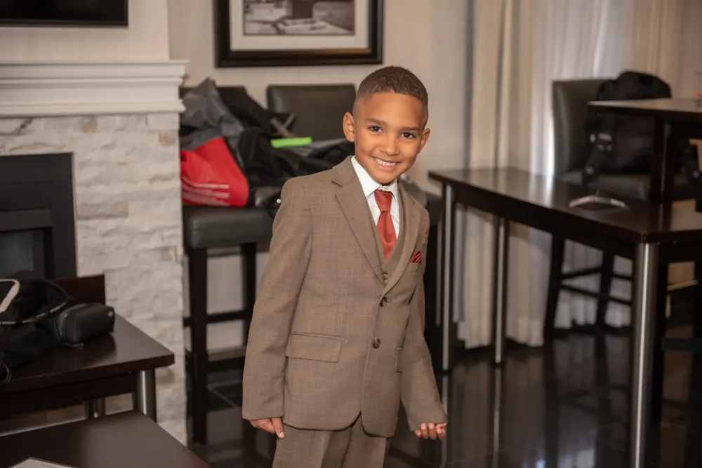 A young boy in a brown suit, white shirt, and red tie smiles in a hotel lobby with luggage and tables nearby.
