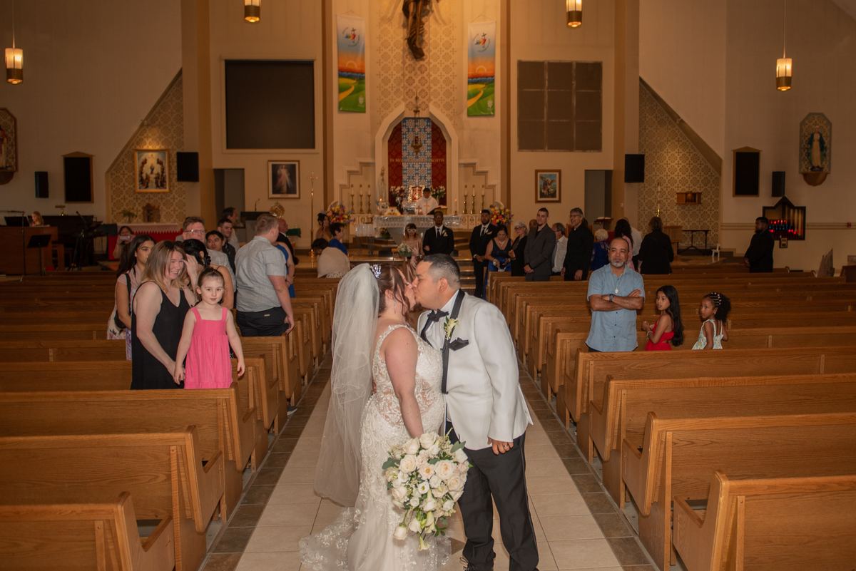Bride and groom kiss at an altar surrounded by guests in wooden pews, illuminated by hanging lights.