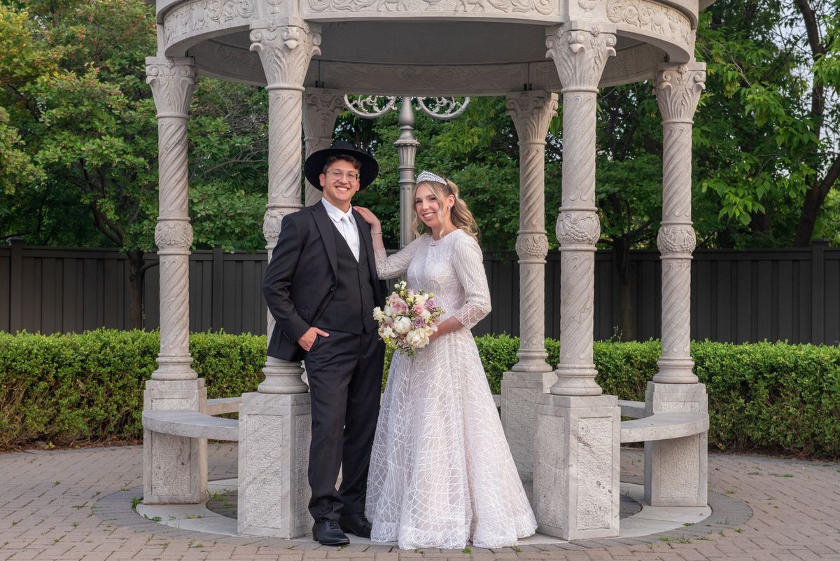 A Jewish couple smiles beneath an ornate stone gazebo in a garden, illuminated by soft daylight and intricate carvings.