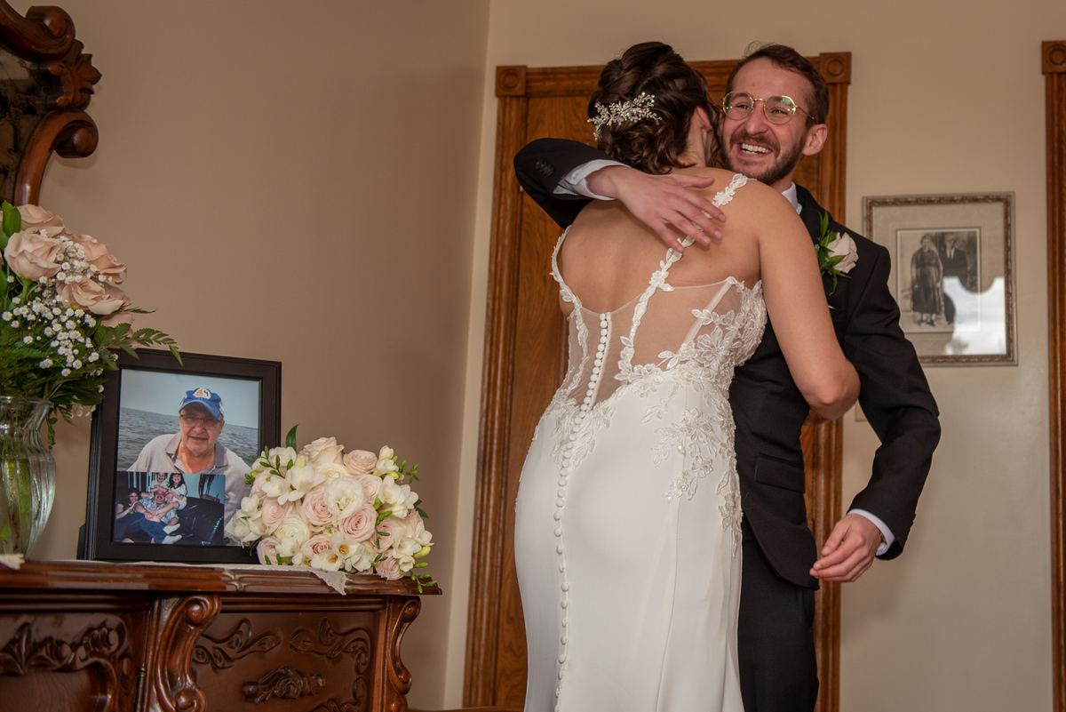 Bride and groom embrace warmly near a dresser with a framed photo of an older man, honoring a memory.