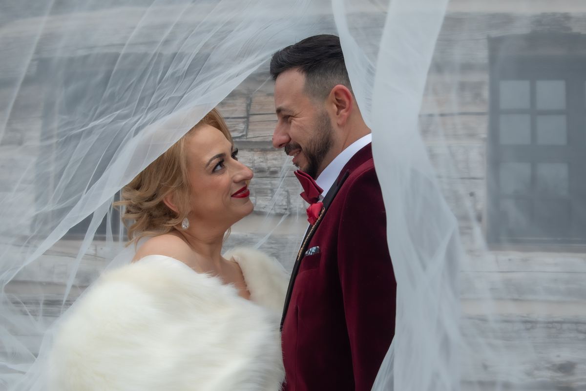 Bride and groom smile at each other, their faces framed by the bride's veil in front of a rustic wooden building.