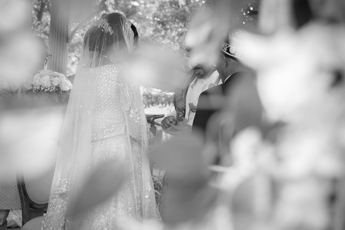 Bride in white gown and veil exchanges rings with groom through blurred foliage, illuminated by natural light.