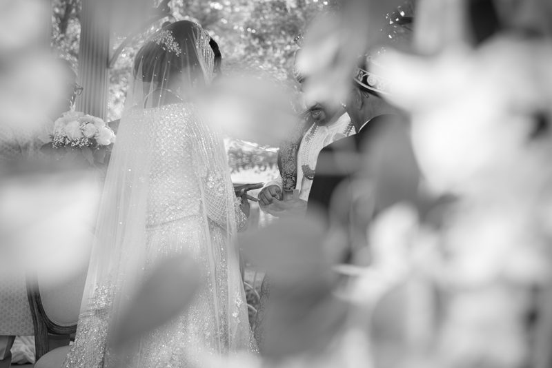 Bride in white gown and veil exchanges rings with groom through blurred foliage, illuminated by natural light.