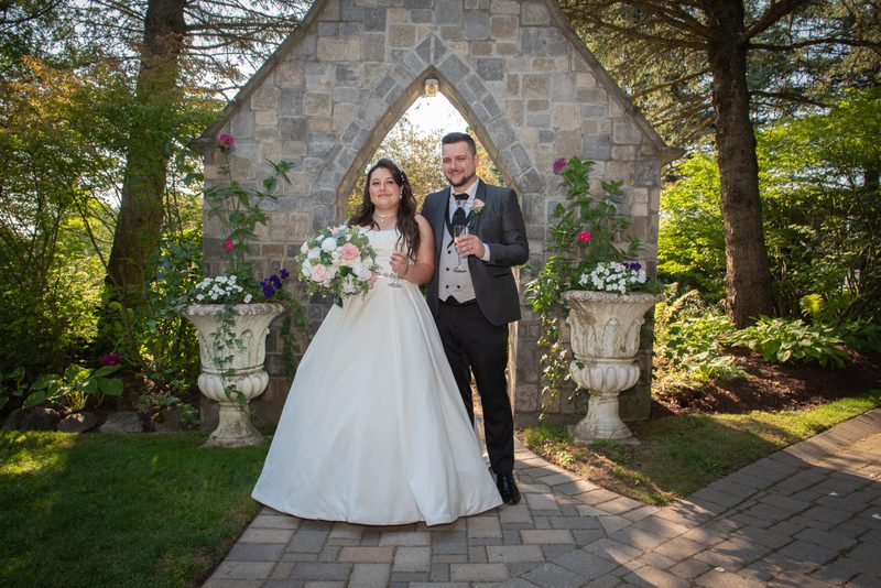A bride and groom smile under a stone archway, holding champagne flutes near a sunlit path surrounded by greenery.