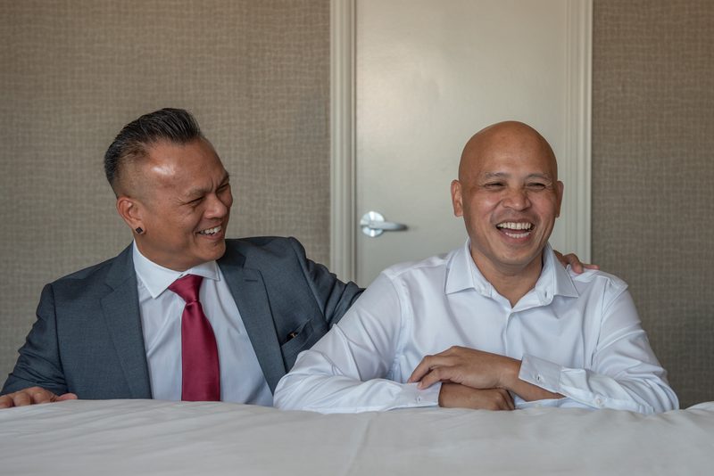 Two men in a hotel room, one in a gray suit and red tie laughing with an arm around his companion in a white shirt.